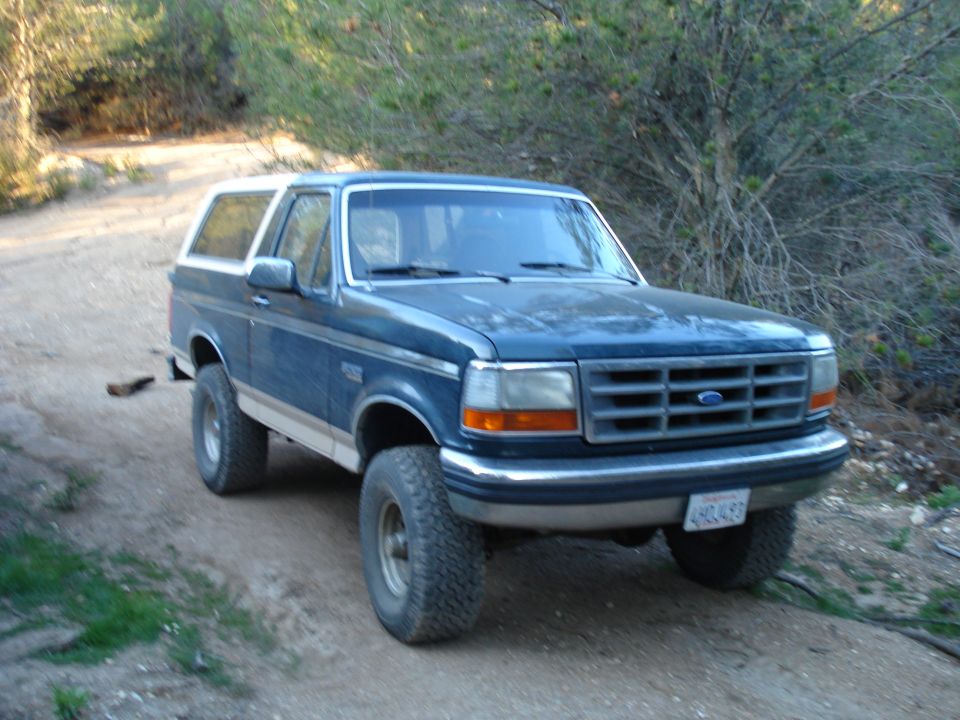 Blue Ford Bronco with white top and tan lower trim, parked on a dirt road