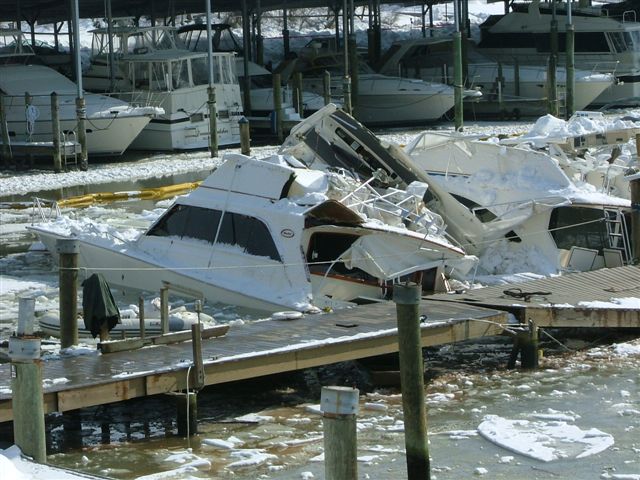 Crushed and half-sunk cabin cruisers piled at an icy marina beside a wooden dock