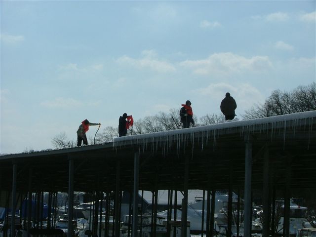 Four people in orange life vests stand on a covered dock with icicles hanging from the edge. Boats are visible below