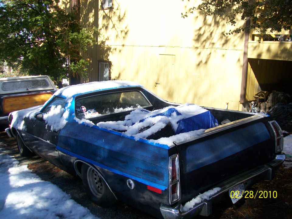 Black Ford Ranchero with blue painted bed, covered in snow, parked next to a pickup truck