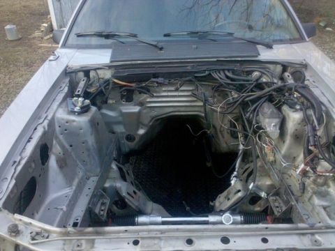 1986 Ford Mustang - The engine bay of a silver 1986 Ford Mustang is shown with the engine removed, revealing exposed wiring and the steering rack.