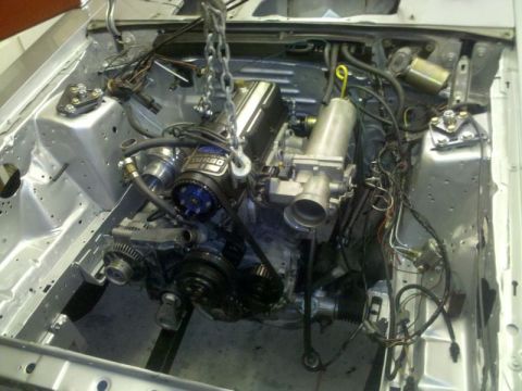 1986 Ford Mustang - Turbocharged inline-four engine lowered into a freshly painted bare silver engine bay by a hoist chain, with a blue cam gear, exposed timin…
