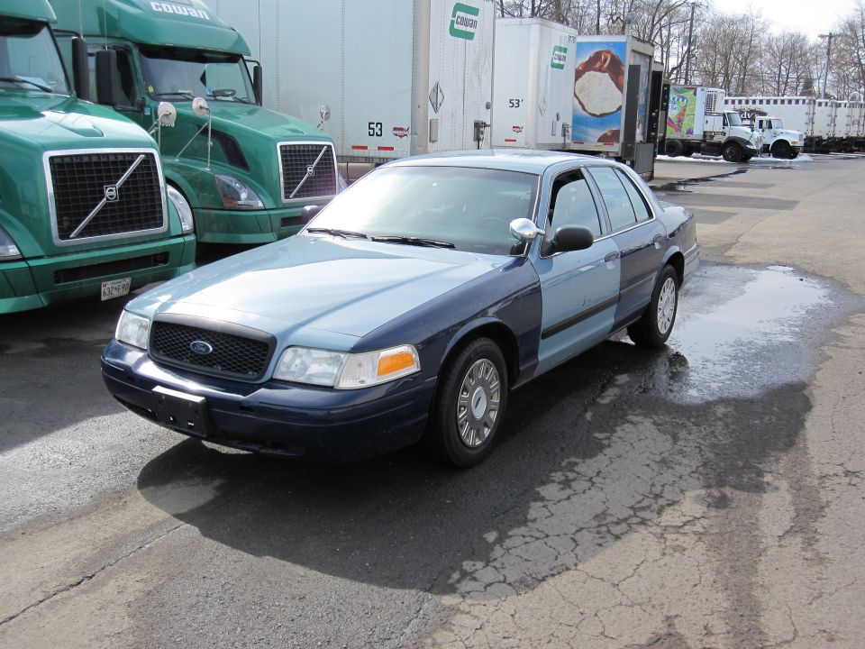 Two-tone blue Ford Crown Victoria, front 3/4 view, parked on cracked asphalt with puddles