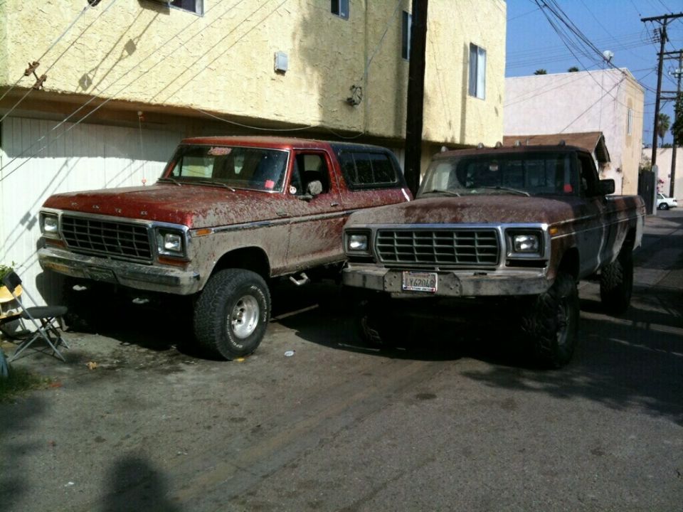 Two weathered Ford trucks parked side-by-side. The one on the left is a red and tan Bronco, the one on the right is a brown pickup. Both have lifted suspensions