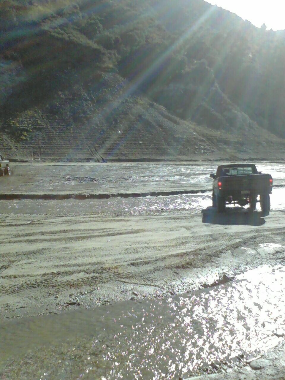 Pickup truck driving on a muddy, wet surface with a large hill in the background. Another vehicle is partially visible to the left