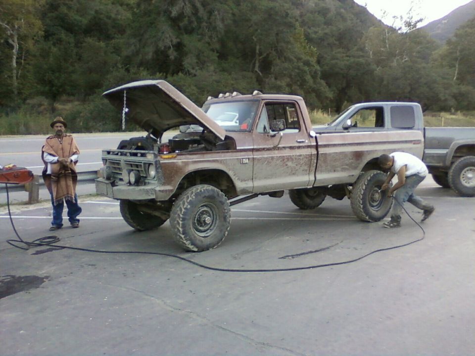 Muddy brown Ford pickup jacked up, hood open, man working on front wheel on roadside