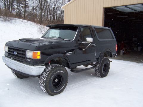 1988 Ford Bronco - This black Ford Bronco, likely from the 1987-1991 generation, is equipped with a hood scoop and aggressive tires for snowy conditions.