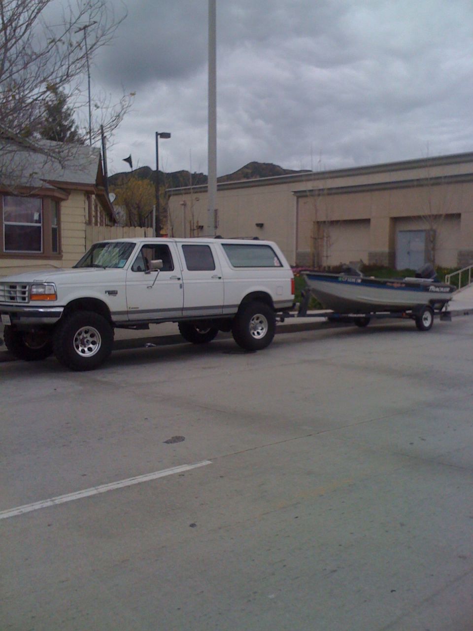 White 1990s Ford Bronco SUV towing a small boat on a trailer, parked on a street