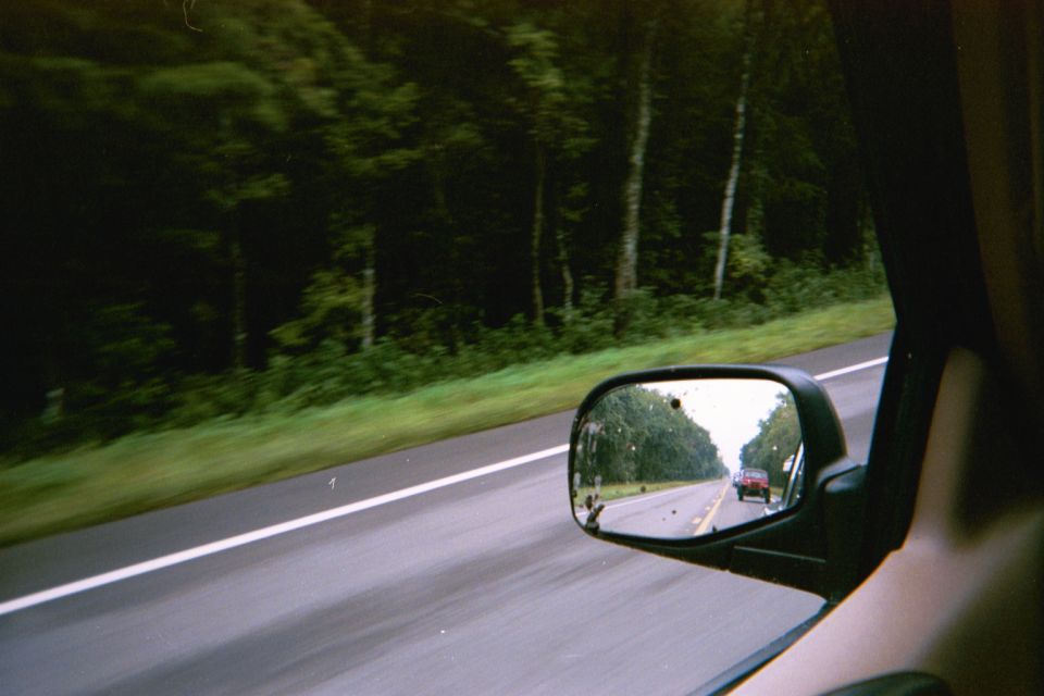 Red car seen in side mirror on a highway with trees lining the road