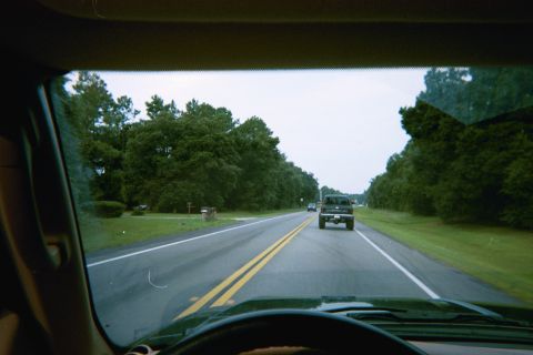 1985 Ford Bronco - A black pickup truck is seen from inside another vehicle driving down a two-lane road lined with trees.