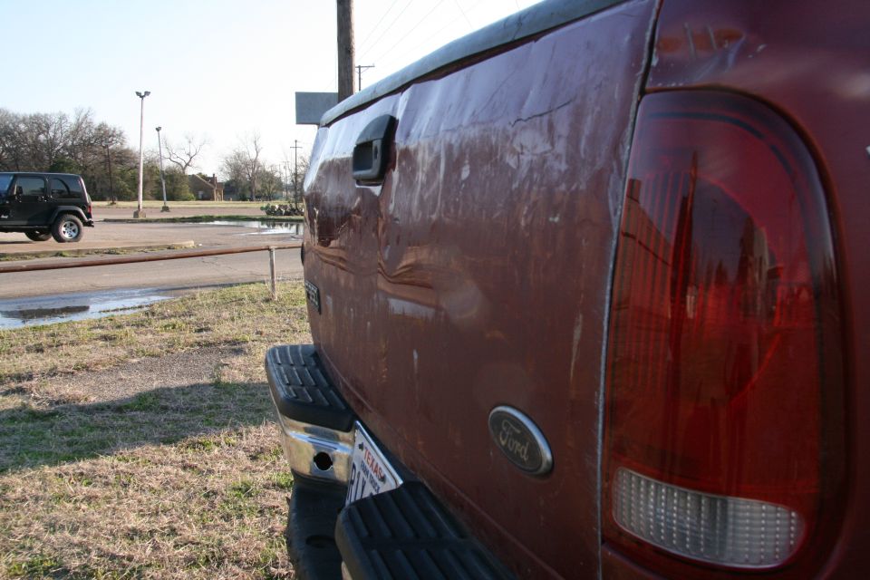 Red Ford pickup truck rear quarter panel with dent, showing Ford oval badge and taillight. Another black Jeep is visible in the background