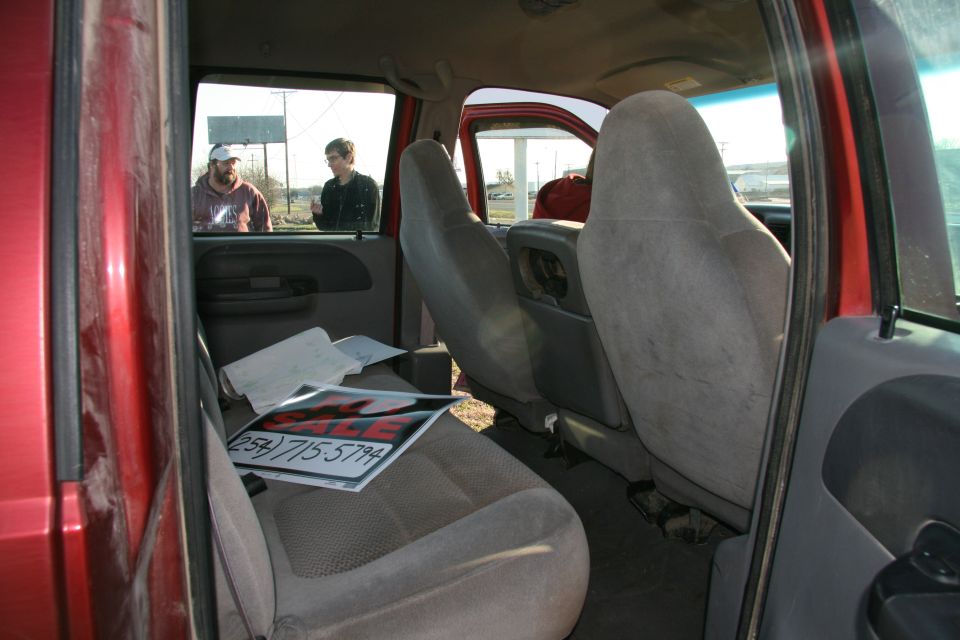 Red truck interior with cloth seats, a "FOR SALE" sign on the passenger seat, and two men visible through the window
