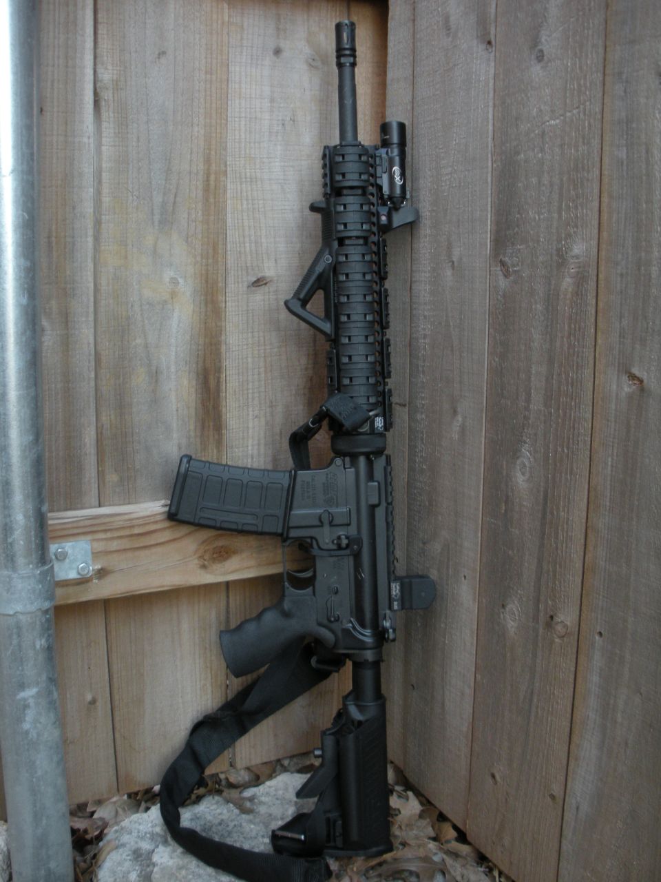 AR-15 style rifle with a flashlight mounted on the rail, leaning against a wooden fence