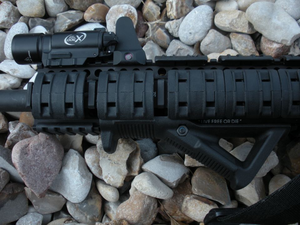 Black rifle with a tactical flashlight and foregrip, resting on rocks. Text reads "LIVE FREE OR DIE"