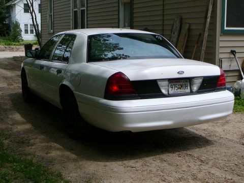 2003 Ford Crown Victoria - Rear view of a white 2003 Ford Crown Victoria sedan parked on a dirt driveway next to a building.