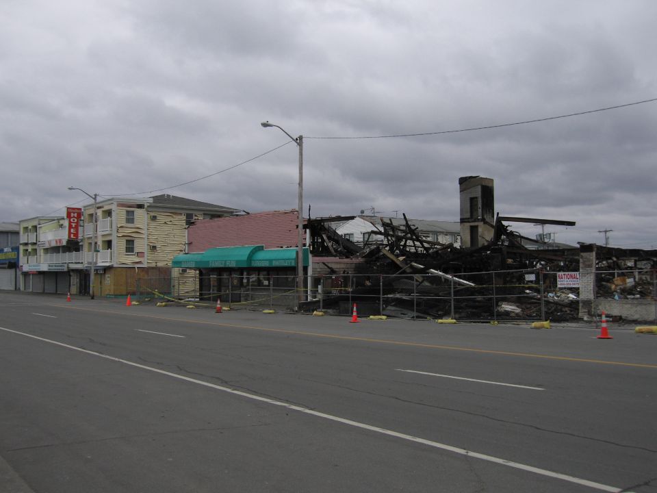 Burned-out building with charred debris behind a fence, orange traffic cones on the roadside