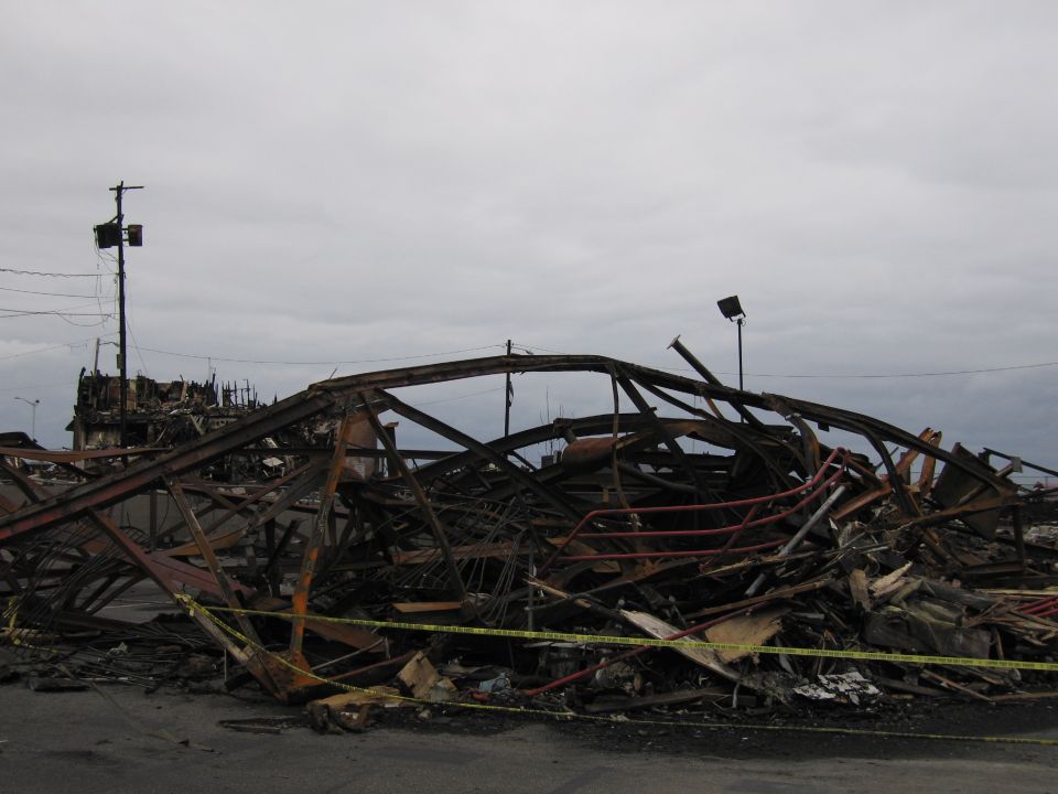 Debris pile from a fire, showing twisted metal beams and charred wood under a cloudy sky. Yellow caution tape is visible