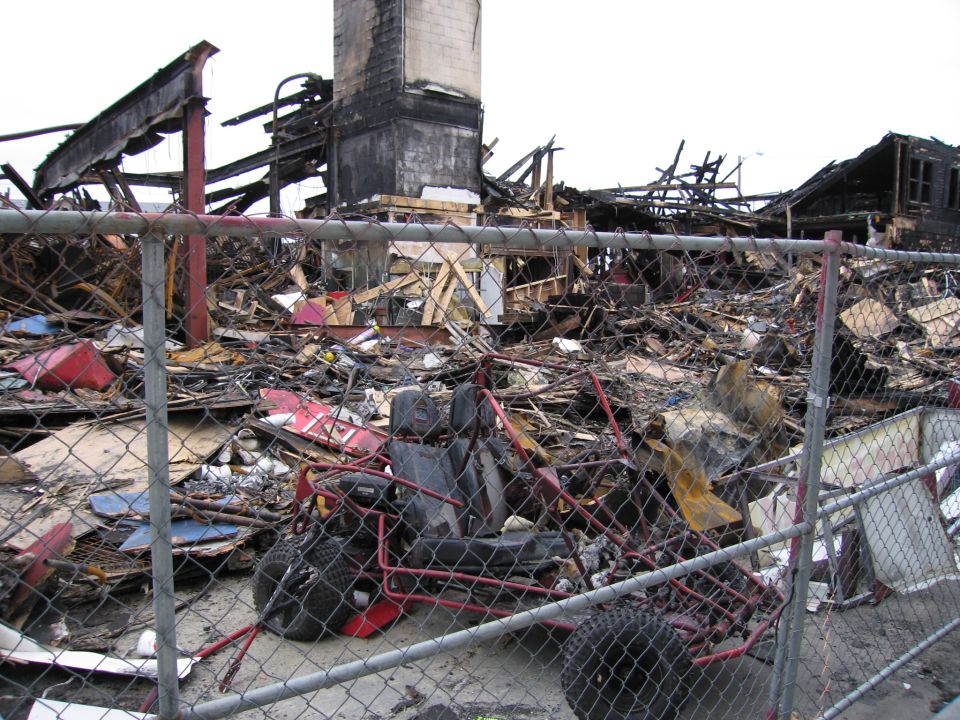 Debris from a fire, including a small red go-kart, behind a chain-link fence