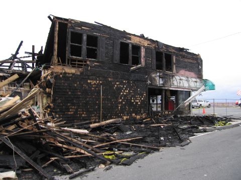 2001 Ford Crown Victoria - A building is heavily damaged by fire, with debris scattered on the street and a white pickup truck visible in the background.