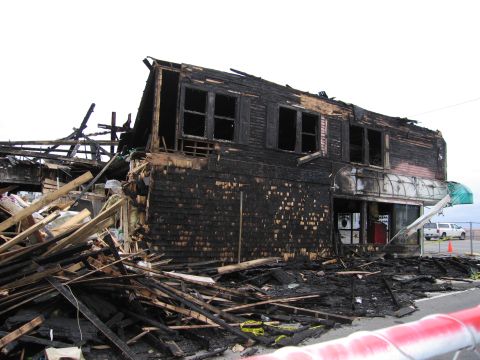 2001 Ford Crown Victoria - A building heavily damaged by fire, with debris scattered across the ground and a white pickup truck parked nearby.