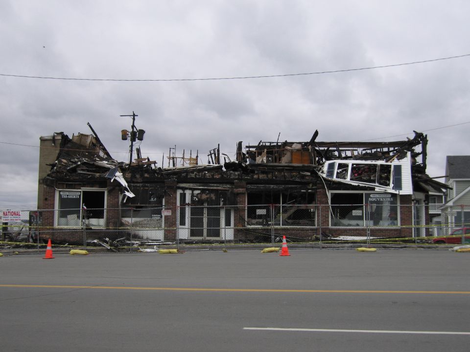 Burned-out brick building with collapsed roof and debris, behind a fence