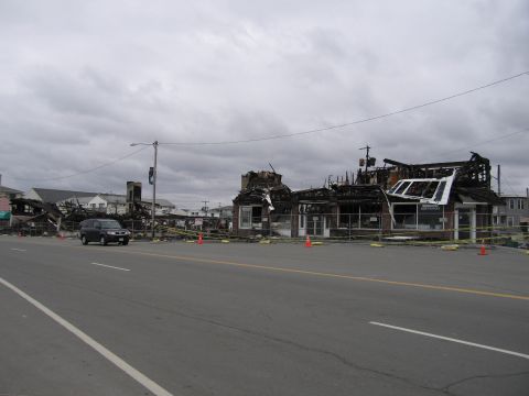 2001 Ford Crown Victoria - A dark SUV is seen driving past a street lined with buildings that have been damaged by fire, with yellow caution tape visible.