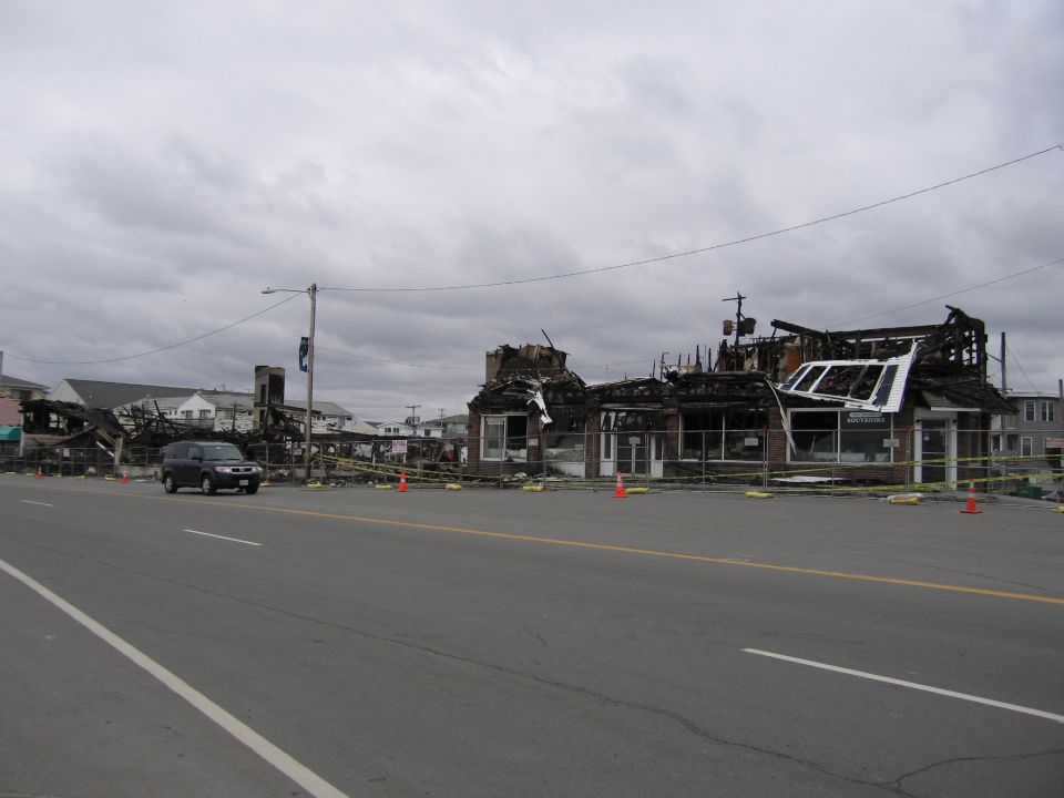 Dark SUV drives past fire-damaged buildings under a cloudy sky