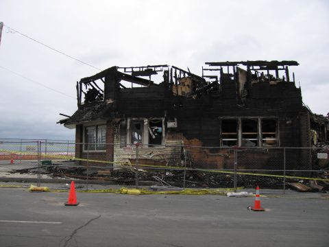2001 Ford Crown Victoria - A building stands heavily damaged by fire, with its roof structure exposed and walls blackened. The scene is cordoned off with fencing and…