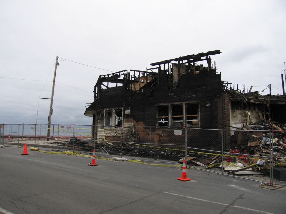 Burned-out house with exposed roof beams, charred walls, and debris piled up behind a chain-link fence