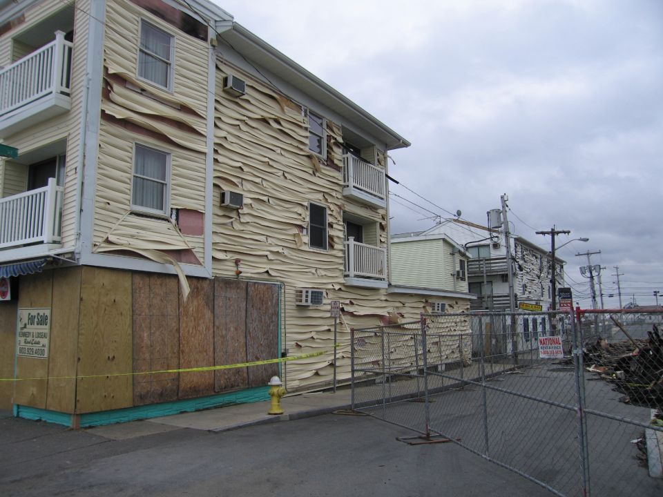 Buildings with siding peeling away, boarded up windows, and a fire hydrant. Chain-link fence in foreground