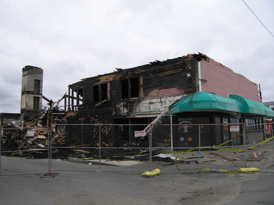 Building heavily damaged by fire, with charred walls, collapsed roof, and debris scattered around. Fencing surrounds the area