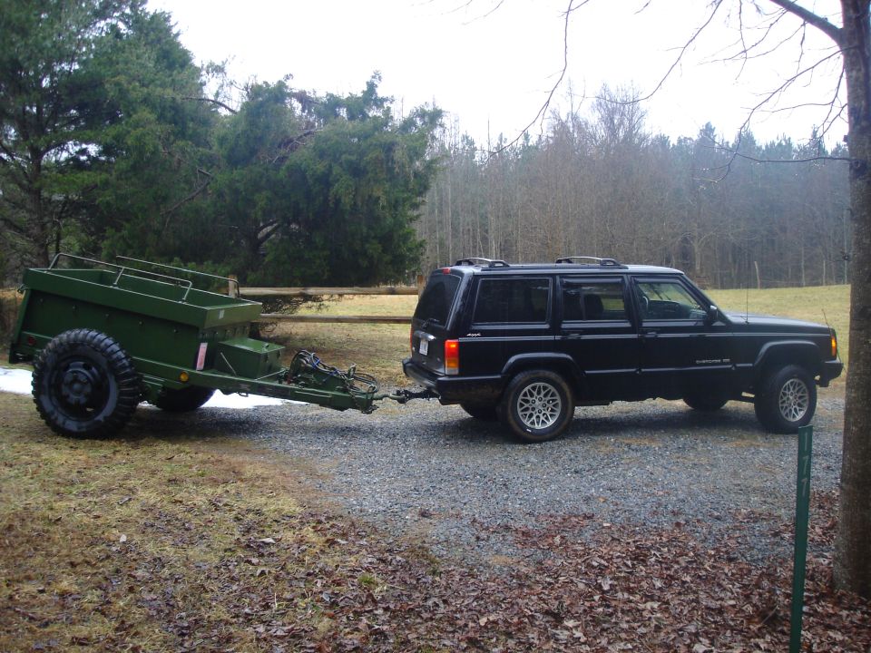 Black Jeep Cherokee with silver rims towing a green utility trailer on a gravel road