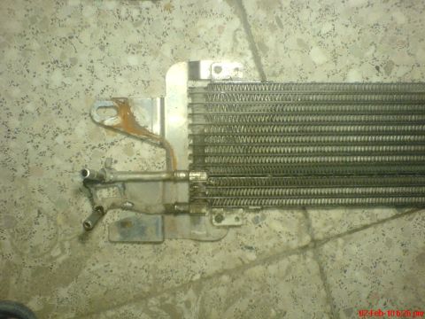 1998 Ford Crown Victoria - A transmission cooler sits on a speckled floor, showing signs of rust and wear on its mounting bracket.