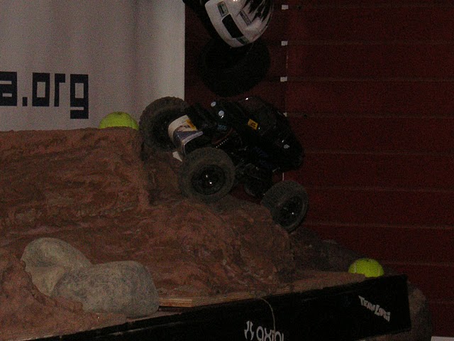 Black remote control rock crawler climbing a dirt mound. A helmet is visible above