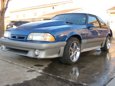1990 Ford Mustang - A blue 1990 Ford Mustang with a prominent cowl induction hood is viewed from a three-quarter front angle on a wet surface.
