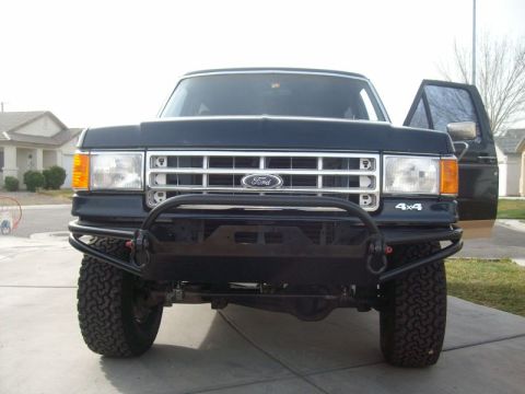 1993 Ford Bronco - Head-on view of a lifted dark blue Ford full-size SUV on a residential driveway, wearing a custom black tube front bumper with a pre-runner…
