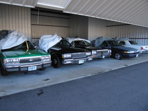 1991 Ford Crown Victoria LTD - Several cars are parked under a metal roof, with the front of a green Ford Crown Victoria clearly visible.