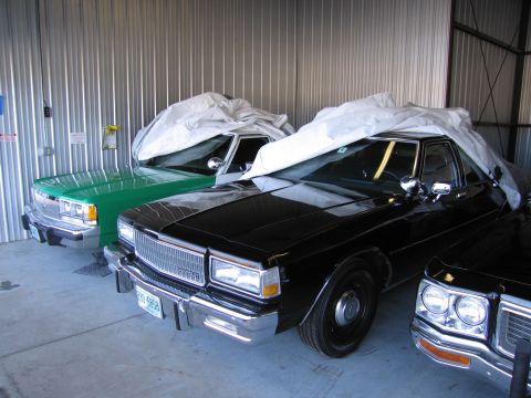 1991 Ford LTD Crown Victoria - Two sedans are parked inside a metal building, one black and one green, both partially covered.