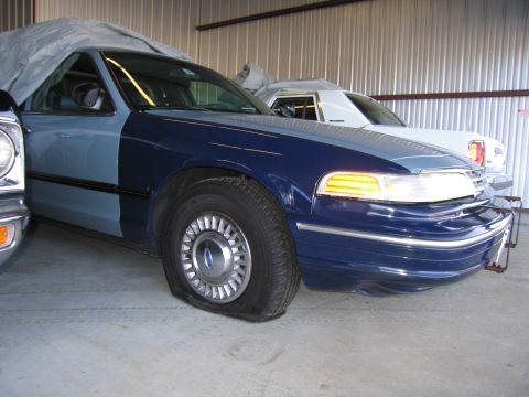 1991 Ford LTD Crown Victoria - A blue Ford Crown Victoria with a flat tire is parked indoors next to another covered vehicle.