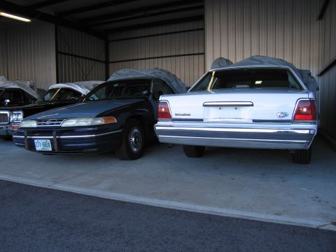 1991 Ford LTD Crown Victoria - Two Ford Crown Victorias are parked inside a garage, one white and one blue, both covered.
