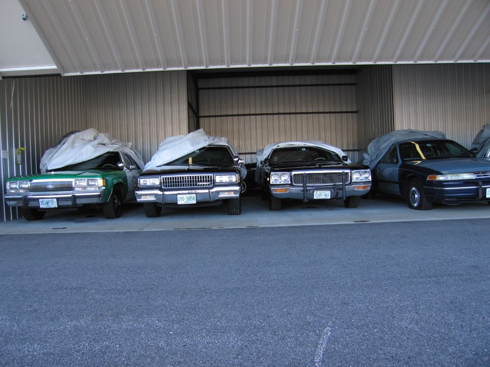 Four cars covered with tarps are parked in a garage. The leftmost car is green