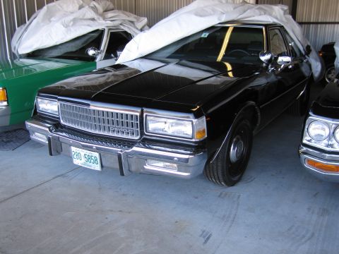 1991 Ford LTD Crown Victoria - A black sedan, partially covered, is parked in a garage next to other vehicles.