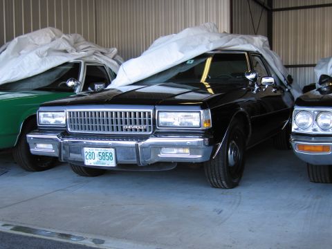 1991 Ford LTD Crown Victoria - A black Chevrolet sedan is parked next to other vehicles, some covered, inside a metal building.