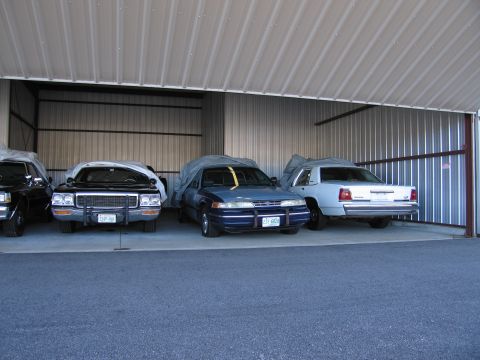 1991 Ford LTD Crown Victoria - Multiple sedans are stored under a metal roof, with two vehicles covered by tarps.