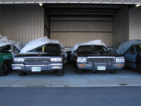 1991 Ford LTD Crown Victoria - Two older sedans are parked side-by-side in a garage, partially covered with protective tarps.