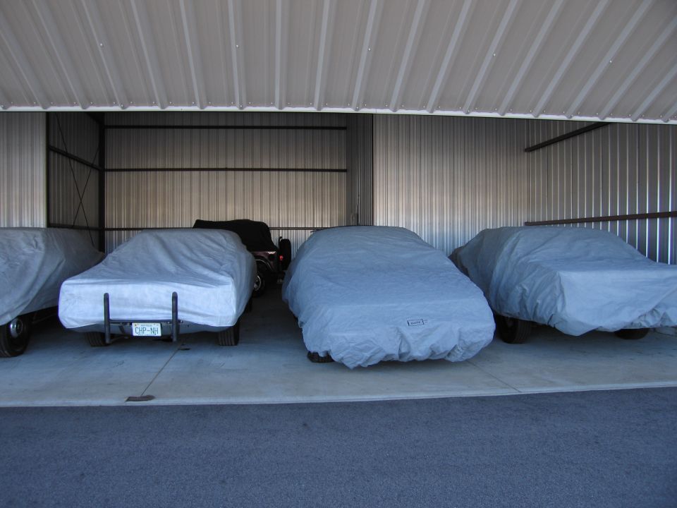 Four cars covered in gray tarps inside a metal storage building. One car's license plate is visible
