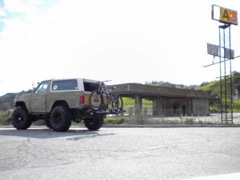 1989 Ford Bronco - A modified 1980s Ford Bronco is parked near an abandoned building with a bicycle mounted on its rear carrier.