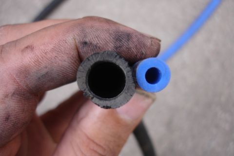 1995 Ford Taurus - A dirty hand holds a worn black hose next to a new blue silicone hose, illustrating a comparison of parts.