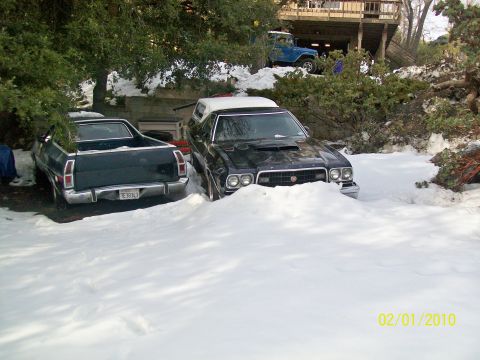1973 Ford Ranchero - Two Ford Rancheros are partially buried in snow, with a blue Jeep on an elevated driveway in the background.