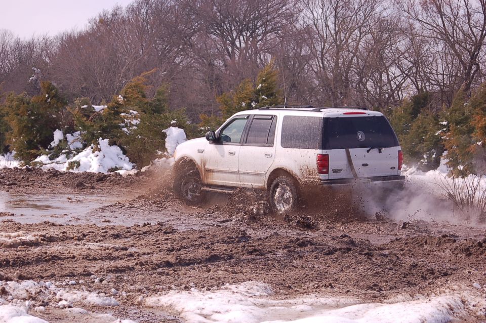 White Ford Expedition driving through a muddy, snowy area. The rear of the vehicle is visible as it kicks up mud